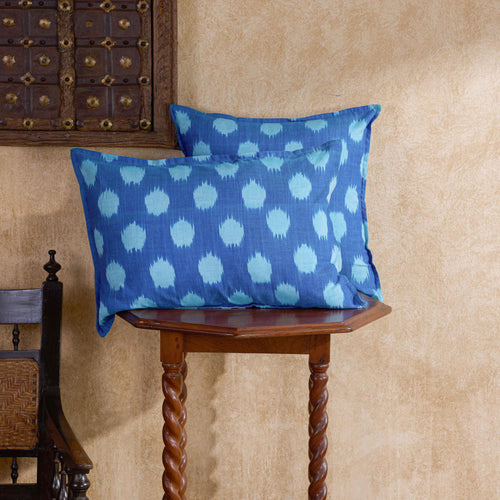 Two blue decorative throw pillows with a light blue ikat dot pattern, displayed on a rustic wooden table against a textured beige wall.